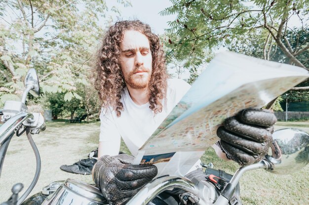 Portret młodego mężczyzny z długimi włosami na motocyklu sprawdzającego ciekawą mapę drogową podczas podróży. Koncepcja podróży. Rękawiczki i niegrzeczne dłonie trzymające mapę nad oldschoolowym motocyklem.