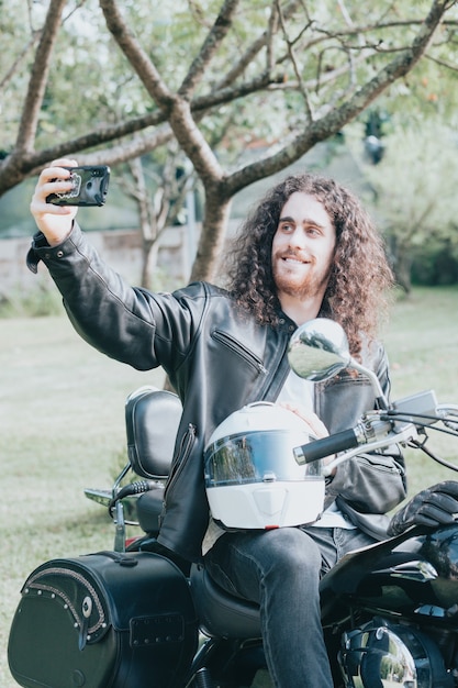 Portret młodego mężczyzny z długimi włosami, starego rowerzysty w skórzanej kurtce na rowerze retro, vintage classic motocykla. pojęcie wolności i stylu, hobby na całe życie. kopiuj przestrzeń, robi selfie