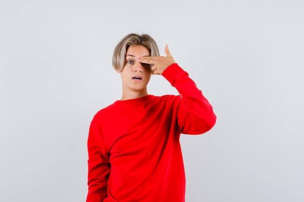Portret młodego nastoletniego chłopca zakrywającego oko palcami w geście pistoletu w czerwonym swetrze i patrzącego na zdziwionego widok z przodu