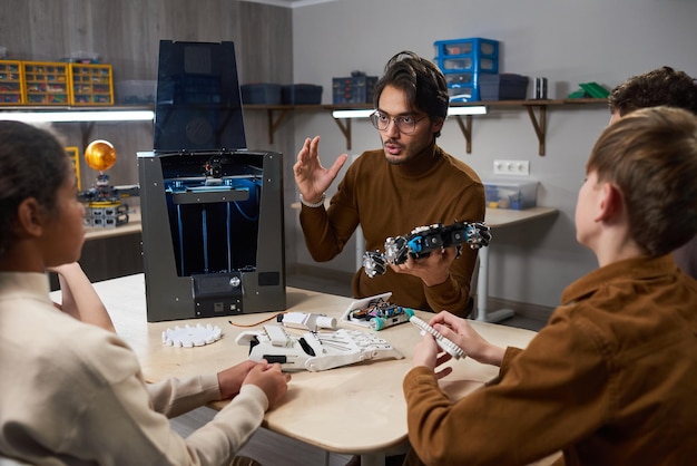 Portret młodego nauczyciela płci męskiej demonstrującego technologię druku 3D zróżnicowanej grupie dzieci na zajęciach z robotyki w szkole