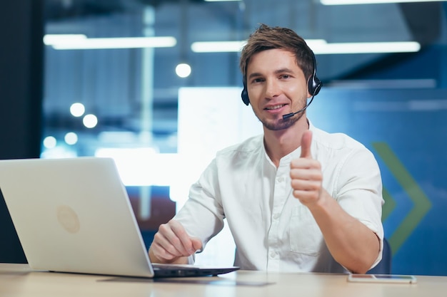 Portret młodego operatora call center, mężczyzny uśmiechającego się i patrzącego w kamerę pracującego z laptopem przy użyciu zestawu słuchawkowego do wideorozmowy