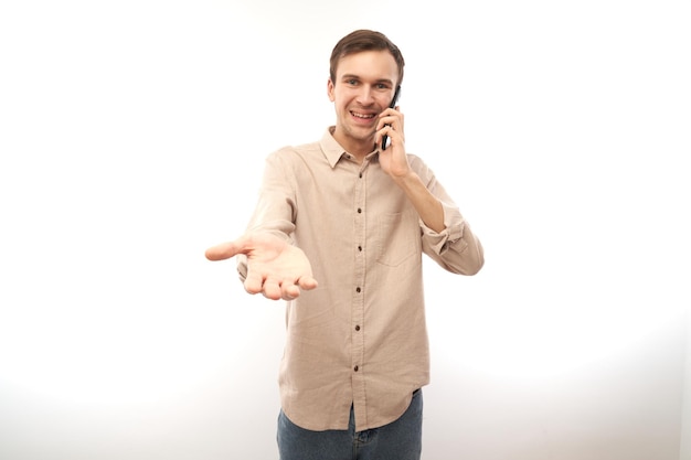Portret młodego przystojnego mężczyzny rasy kaukaskiej używającego telefonu komórkowego i uśmiechającego się, wyciągającego rękę do kamery na białym tle na białym tle studia radosny wyraz twarzy