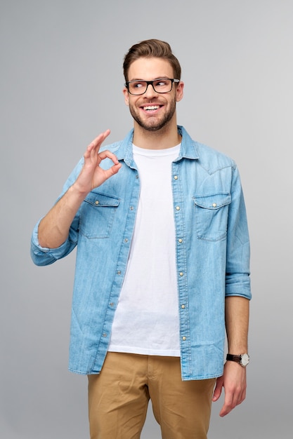 Portret młodego przystojnego mężczyzny w koszuli dżinsy Wyświetlono ok znak gest stojąc nad jasną ścianą