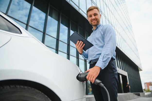 Portret młodego przystojnego mężczyzny w stroju codziennym stojącego przy stacji ładującej Koncepcja ekologicznego samochodu elektrycznego