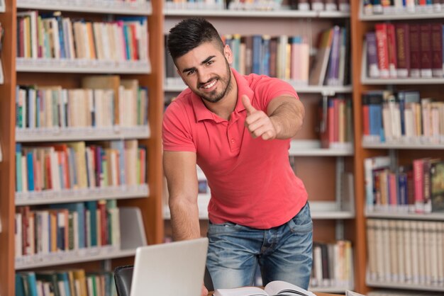 Portret młodego, sprytnego studenta pokazującego kciuki w bibliotece uczelni Płytkiej głębi ostrości