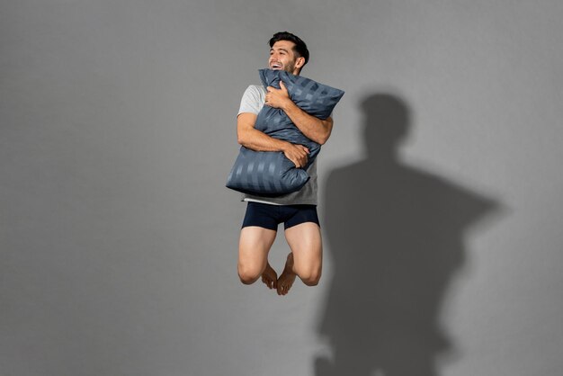 Portret młodego świeżego energicznego mężczyzny noszącego bieliznę nocną trzymającą poduszkę i skaczącego w powietrzu po przebudzeniu z dobrego snu rano, studio strzał w szarym na białym tle