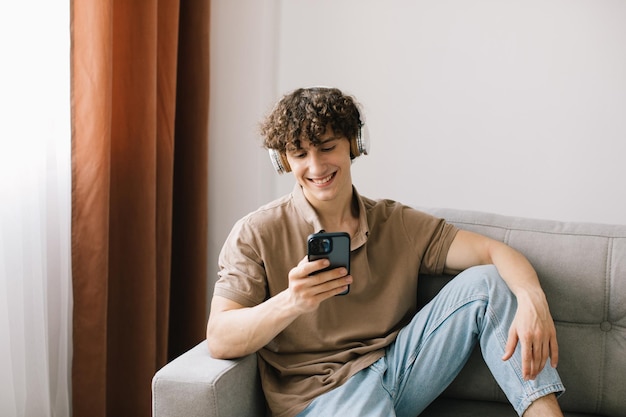 Portret młodego szczęśliwego mężczyzny z kręconymi włosami za pomocą smartfona ze słuchawkami, siedząc na kanapie w salonie i odpoczywając