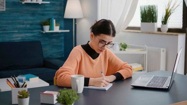 Portret młodego ucznia robiącego notatki w podręczniku w klasie online na laptopie, uczestniczącego w zdalnym spotkaniu z nauczycielem. Kobieta pisze informacje o uniwersytecie na notebooku do nauki.