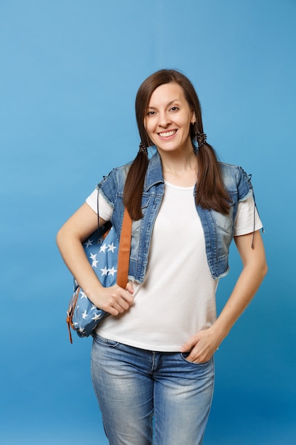 Portret młodego uśmiechniętego studenta ładnej kobiety w biały t-shirt, dżinsowe ubrania z plecakiem, trzymając rękę w kieszeni na białym tle na niebieskim tle. Edukacja w szkole średniej. Skopiuj miejsce na reklamę.