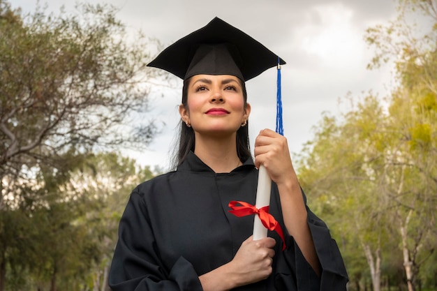 Portret młodej absolwentki szkoły w czapce i sukni, trzymającej dyplom na zewnątrz