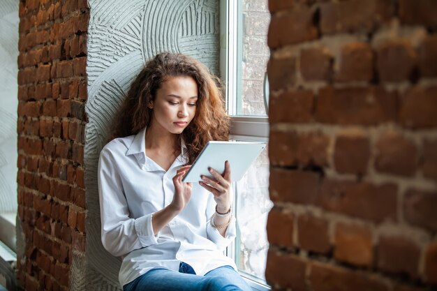 Zdjęcie portret młodej afrykańskiej kobiety siedzącej w biurze za pomocą cyfrowego tabletu. wesoła afrykańska kobieta korzystająca z tabletu