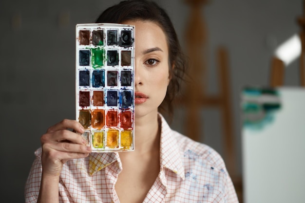 Portret młodej artystki obejmującej twarz farbami akwarelowymi. Kreatywna malarka patrząca poważnie w kamerę. Koncepcja kreatywności i indywidualności