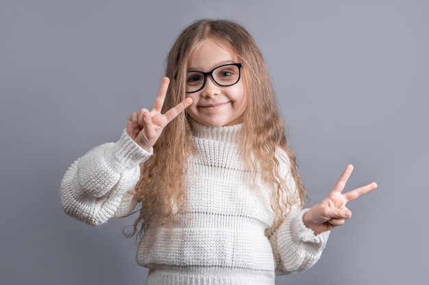 Portret młodej atrakcyjnej dziewczynki z blond włosami w swetrze przedstawiającym znak v, znak pokoju, gest zwycięstwa na obu rękach na szarym tle studio.