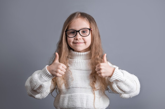 Portret młodej atrakcyjnej dziewczynki z długimi blond włosami w białym swetrze uśmiecha się pokazuje kciuki do góry, jak śpiewać Miejsce dla tekstu. Skopiuj miejsce.