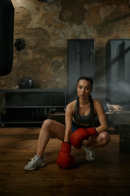 Portret młodej atrakcyjnej kobiety boksera lub kickboxera w dresach i rękawicach do wykrawania w siłowni na poddaszu. Sztuki walki i kobieca koncepcja