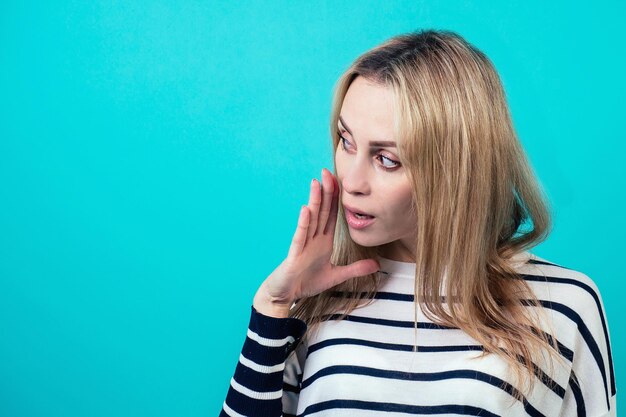 Zdjęcie portret młodej atrakcyjnej kobiety o blond włosach i makijażu szepcze tajną tajemnicę w studio na niebieskim tle. pojęcie plotek i poufności.