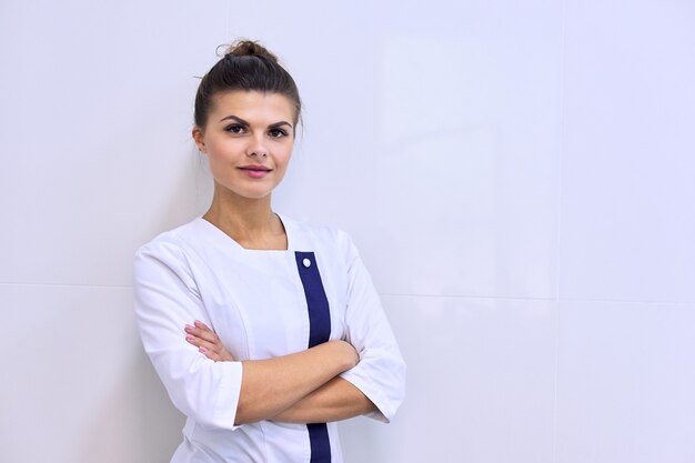 Zdjęcie portret młodej atrakcyjnej kobiety pewnie w mundurze medycznym z rękami skrzyżowanymi, tło kliniki światła ściany, miejsce. farmaceuta, pielęgniarka, kosmetolog, specjalista, naukowiec