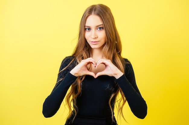 Zdjęcie portret młodej atrakcyjnej kobiety z długimi włosami rękami gestykuluje serce w studio na żółtym tle. koncepcja walentynek i wyznania miłości