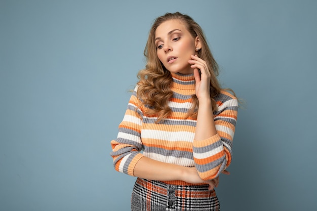Portret młodej atrakcyjnej pięknej przemyślanej seksownej blond osoby płci żeńskiej noszącej stylowy kolorowy sweter w paski na białym tle na niebieskim tle z kopią przestrzeni i myślenia