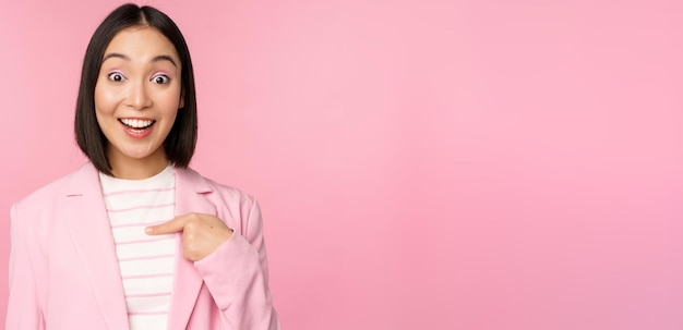 Portret młodej azjatyckiej bizneswoman ze zdziwionym podekscytowanym wyrazem twarzy, wskazując palcem na siebie stojącą w garniturze na różowym tle