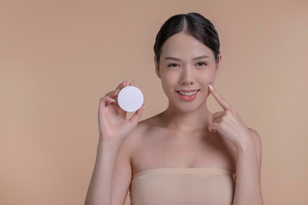 Portret młodej azjatyckiej kobiety uśmiechającej się, trzymającej makietę produktu do reklamy tekstowej, umieszczającej beżowe tło Koncepcja opieki zdrowotnej dla produktu do pielęgnacji skóry dla reklamy