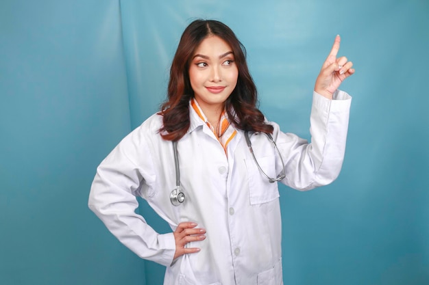 Portret młodej azjatyckiej lekarki, która jest lekarzem, uśmiecha się i wskazuje w górę na kopię odizolowaną na niebieskim tle