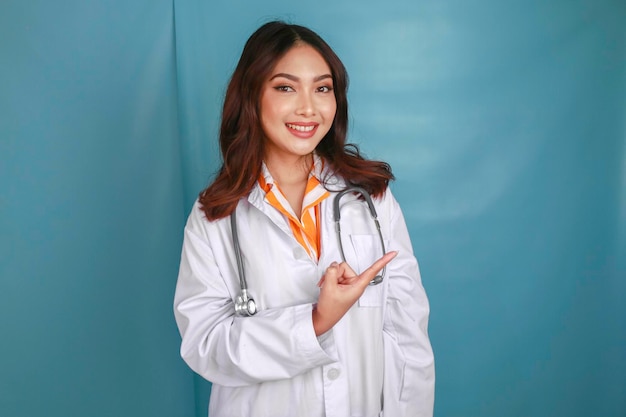 Portret młodej azjatyckiej lekarki, lekarza, który uśmiecha się i wskazuje na kopię miejsca odizolowaną na niebieskim tle