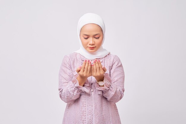 Portret młodej azjatyckiej muzułmanki noszącej hidżab i fioletową sukienkę stojącą z podnoszeniem rąk i modlącą się do boga odizolowanego na białym tle