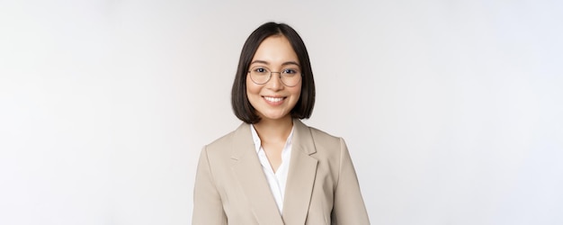Portret młodej azjatyckiej sprzedawczyni w okularach ubrana w beżowy garnitur, uśmiechnięta i patrząca pewnie na białe tło kamery