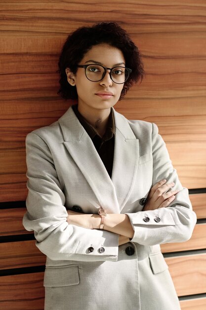 Portret młodej bizneswoman w okularach i białym garniturze stojącej z rękami skrzyżowanymi i spojrzeniem