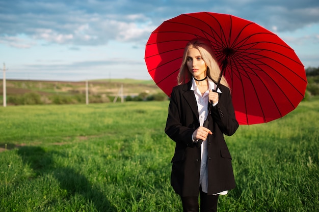 Portret młodej blondynki ładna dziewczyna w czarno-białym garniturze, trzymając czerwony parasol na tle zachmurzonego nieba i zielonego pola.