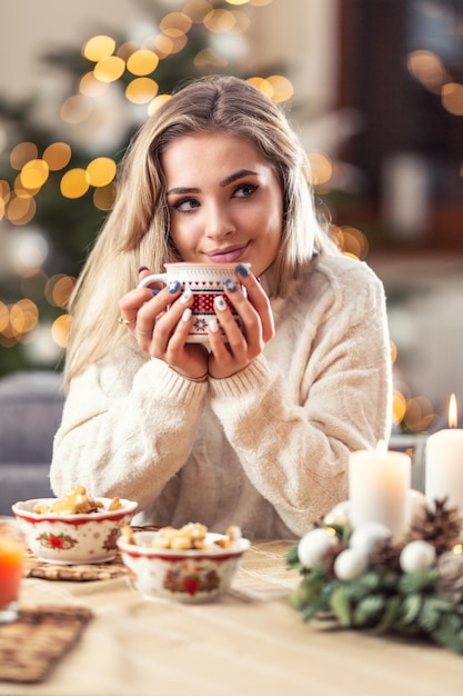 Portret młodej blondynki siedzącej przy wigilijnym stole przed choinką, pijącej gorącą herbatę lub poncz