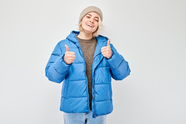 Portret młodej blondynki w zimowym stroju uśmiechającej się radośnie pokazującej gest kciuka w górę izolowany na białym tle studia. Zatwierdza dobry wybór, właściwą decyzję