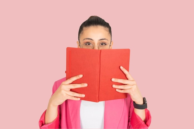 portret młodej brunetki, białej kobiety ukrywającej się za otwartą książką, okularami, formalną różową kurtką