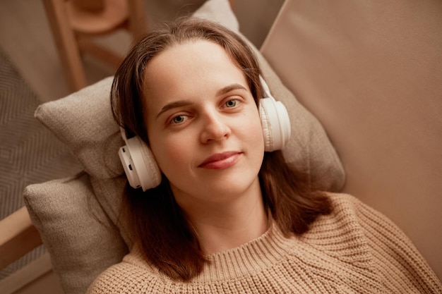 Portret młodej dorosłej białej kobiety o brązowych włosach i zrelaksowanej twarzy, patrzącej na kamerę, słuchającej muzyki przez słuchawki podczas leżenia na sofie