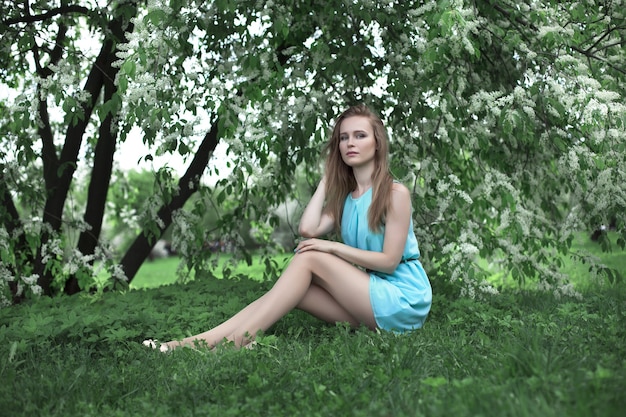 Portret młodej dziewczyny blondynka w niebieskiej sukience. ona siedzi na trawie. drzewa w kwiatach.