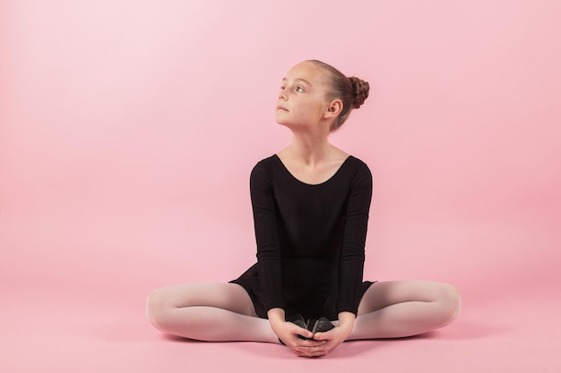 Portret młodej dziewczyny piękne dziecko baleriny praktykujących ćwiczenia rozciągające balet na sobie czarną sukienkę tutu pozowanie Studio z jasnoróżowym tłem.