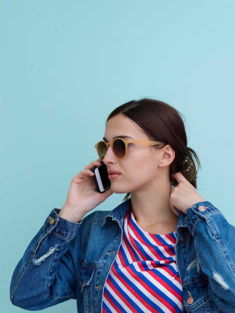 Portret młodej dziewczyny rozmawiającej przez telefon stojąc przed niebieskim tłem. Modelka w okularach przeciwsłonecznych reprezentująca nowoczesną koncepcję mody i technologii