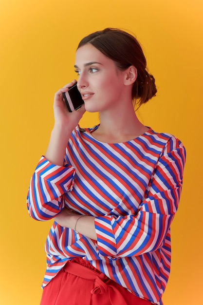 Portret młodej dziewczyny rozmawiającej przez telefon stojąc przed żółtym tłem. Modelka reprezentująca nowoczesną koncepcję mody i technologii