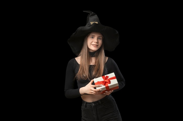 Portret młodej dziewczyny w czarnych ubraniach w spiczastym kapeluszu wiedźmy z prezentem w rękach w izolacji na czarnym tle Halloween