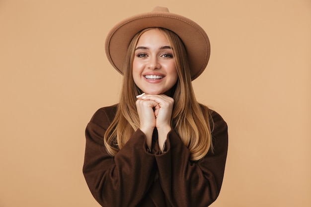 Portret młodej dziewczyny w kapeluszu i płaszczu uśmiechający się na beżowym tle