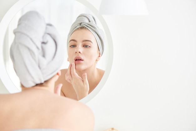 Portret młodej dziewczyny z ręcznikiem na głowie w białej łazience wygląda i dotyka jej twarzy w lustrze i cieszy się młodością i nawodnieniem Naturalna pielęgnacja domowa pielęgnacja skóry problematycznej