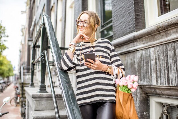 Portret młodej eleganckiej kobiety stojącej na schodach luksusowego domu mieszkalnego w Amsterdamie ze smartfonem