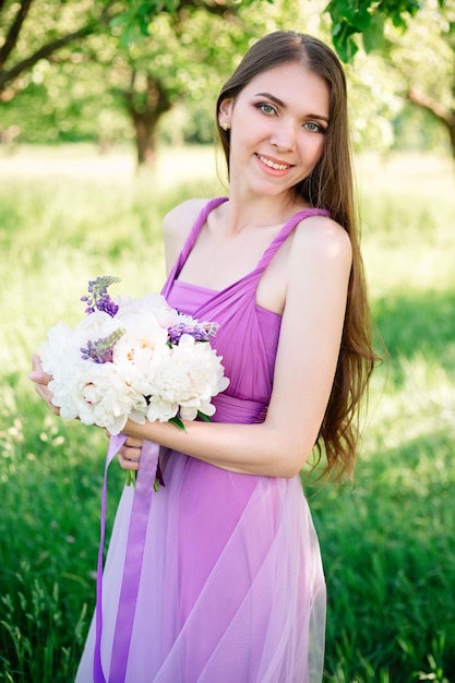 Portret młodej kobiety 20s 30s w jasnofioletowej sukience z bukietem piwonii w dłoniach.