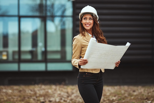 Zdjęcie portret młodej kobiety inżynier z białym kaskiem i plany w jej ręce na budowie.