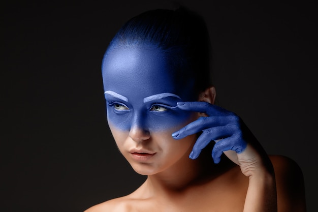 Portret młodej kobiety, która pozuje pokryta niebieską farbą w studio na czarnym tle. dłoń dziewczyny wokół twarzy i pomalowana na niebiesko