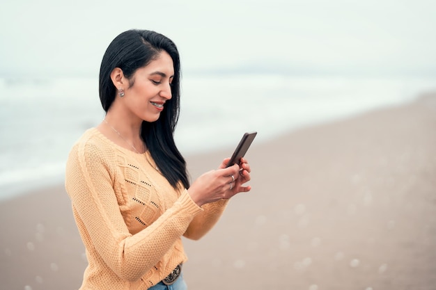 Portret młodej kobiety latynoskiej na plaży uśmiechający się szczęśliwy patrząc na telefon komórkowy