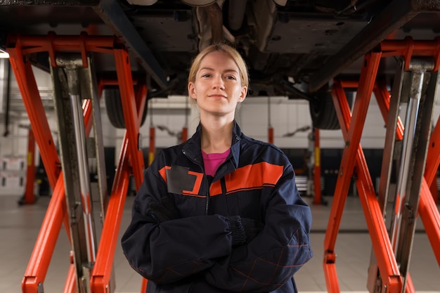 Portret młodej kobiety-mechanika w mundurze, która patrzy w kamerę na tle samochodów naprawianych na stacji paliw