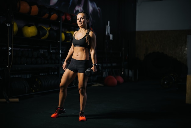 Portret młodej kobiety mięśni z doskonałego ciała atletycznego na sobie czarną odzież sportową, stojąc w nowoczesnej ciemnej siłowni. Pojęcie zdrowego stylu życia.