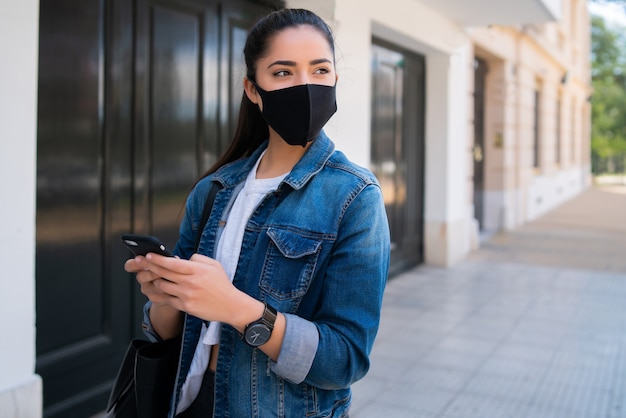 Portret młodej kobiety noszenia maski i korzystania z telefonu komórkowego podczas spaceru na ulicy